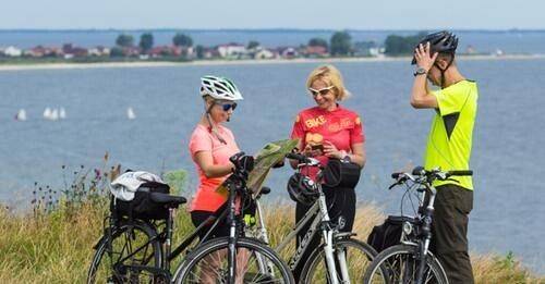 Troje rowerzystów stoi przy rowerach i czyta mapę. W tle akwen wodny. grafika