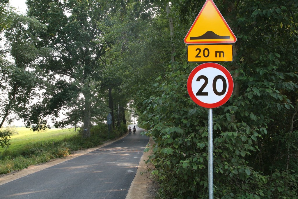 Przebudowany odcinek drogi prowadzący przez pola i las. Na pierwszym planie znak drogowy ostrzegający o progu spowalniającym, który pojawi się na drodze za 20 metrów od miejsca ustawienia znaku oraz znak ograniczenia prędkości do 20 km na godzinę. Zadrzewione pobocze.