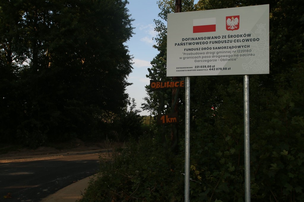 Przebudowany odcinek drogi prowadzący przez las. Przy drodze ustawiona tablica informacyjna z inwestycji, a na niej komunikat o dofinansowaniu z Funduszu Dróg Samorządowych. Na tablicy znajduje się flaga i godło Polski.