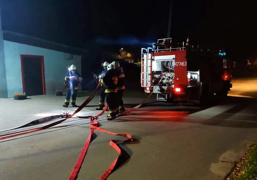 Strażacy przygotowują się do wyjazdu na akcję. Wóz bojowy stoi przed remizą strażacką.