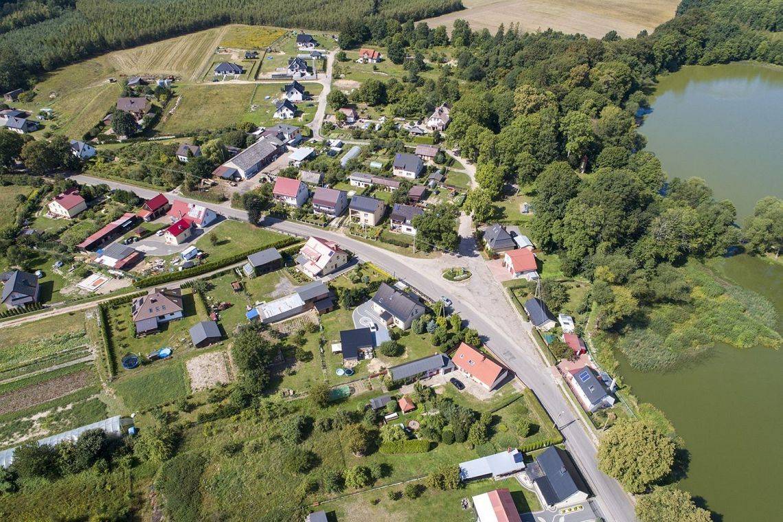 Zdjęcie z lotu ptaka. Widok na drogę w miejscowości Redkowice, zabudowę mieszkaniową, akwen wodny, pola i lasy.
