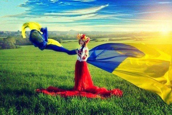 Na łące, w promieniach słońca, stoi kobieta w wianku z kwiatów na głowie i biało-czerwonym stroju. Wokół niej, na wietrze, powiewa niebiesko-żółta wstęga, symbolizująca ukraińską flagę.