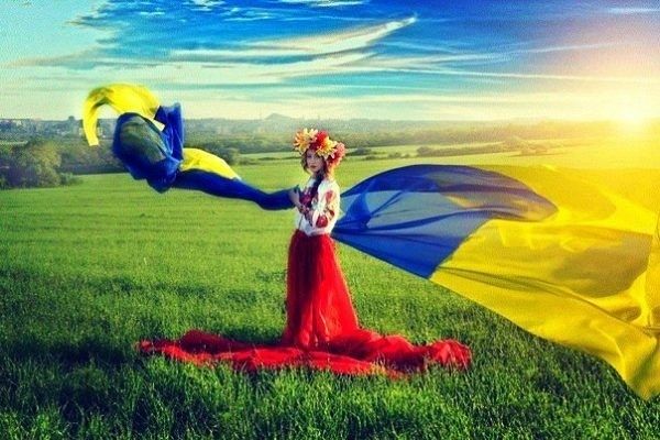 Na łące, w promieniach słońca, stoi kobieta w wianku z kwiatów na głowie i biało-czerwonym stroju. Wokół niej, na wietrze, powiewa niebiesko-żółta wstęga, symbolizująca ukraińską flagę.