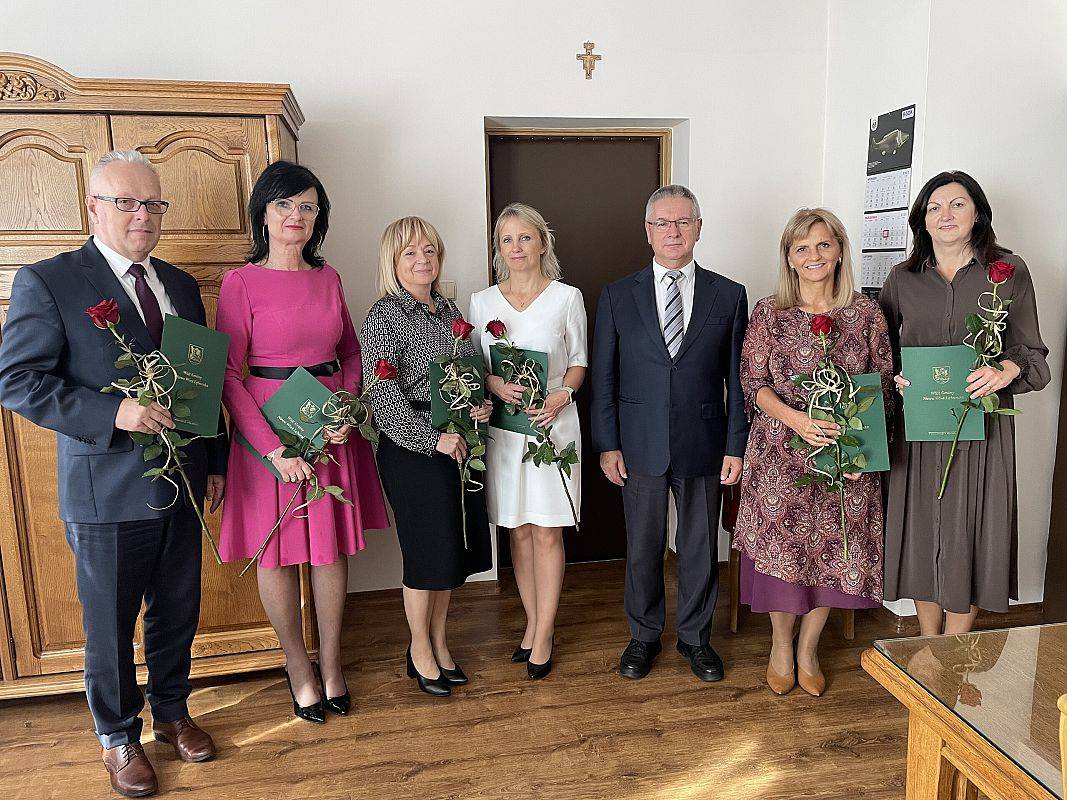 Dyrektorzy szkół (5 osób) i dyrektor przedszkola (1 osoba) z dyplomami i kwiatami stoją w szeregu z wójtem gminy w jego gabinecie.