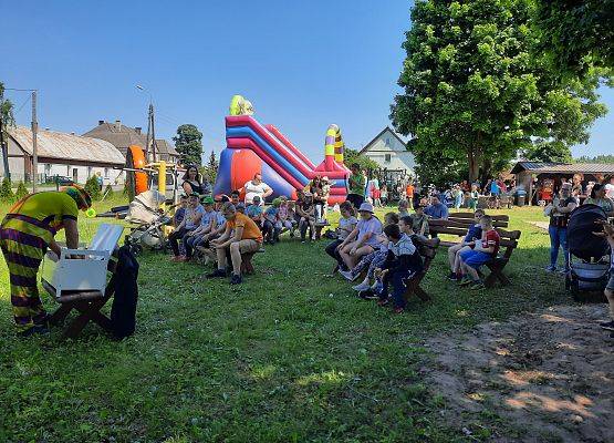 Na terenie zielonym, uczestnicy siedzą pikniku siedzą na ławkach i oglądają pokaz magika, animatora. W tle dmuchane zabawki i zjeżdżalnie