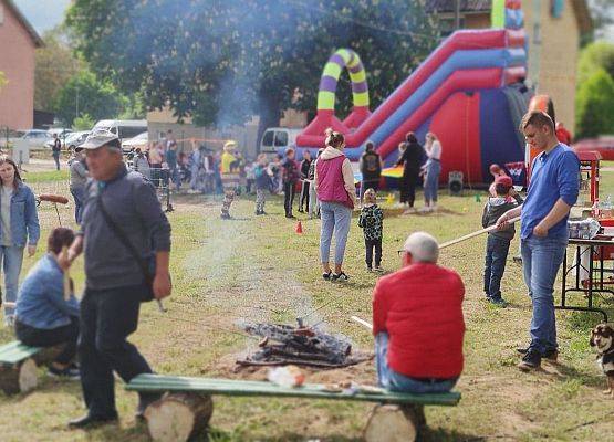 Piknik na terenie zielonym. Dzieci, dorośli, smażą kiełbaski na ognisku, obok stoisko z poczęstunkiem. W oddali dmuchane zabawki i zjeżdżalnie.