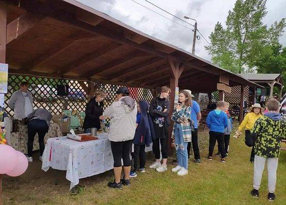 Na terenie zielonym stoły z poczęstunkiem, przekąskami ustawione pod drewnianą wiatą oraz kobiety z koła gospodyń wiejskich obsługujące uczestników pikniku.