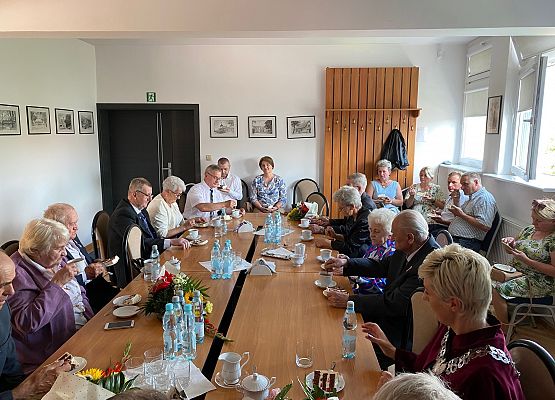 Jubilaci, wójt, zaproszeni goście siedzą przy wspólnym stole przy kawie i torcie.