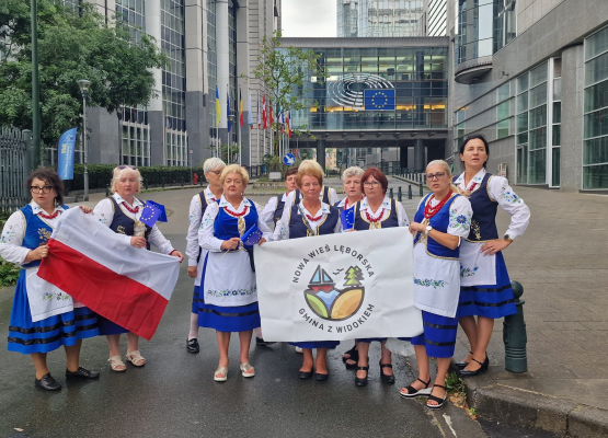 Kobiety w regionalnych strojach pozują do zdjęcia przed budynkiem Parlamentu Europejskiego. Trzymają flagę Polski oraz flagę z logo gminy.