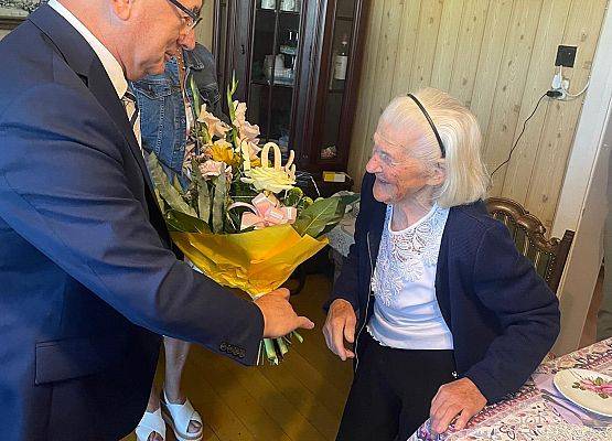 Wójt gminy wręcza bukiet kwiatów jubilatce w jej mieszkaniu.