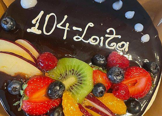 Czekoladowy tort udekorowany różnymi kolorowymi owocami  z napisem 104 lata.