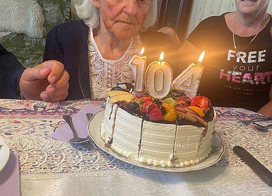 Jubilatka siedzi przy stole, a przed nią tort z zapalonymi świeczkami.