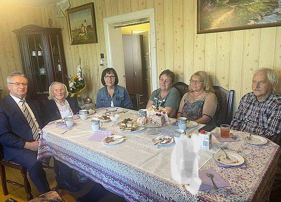 Wójt gminy, jubilatka i zaproszeni goście siedzą przy stole.