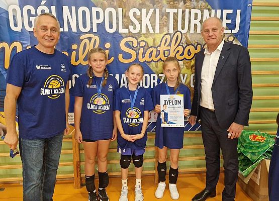 Dwóch dorosłych mężczyzn i trzy zawodniczki z pucharami i dyplomami pozują do zdjęcia w hali sportowej.