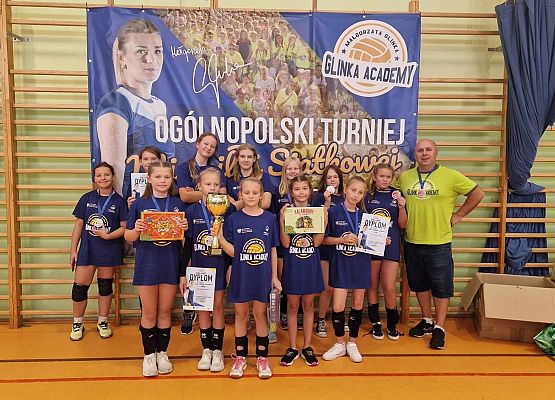 Drużyna zawodniczek piłki siatkowej z trenerem pozują do zdjęcia w hali sportowej.