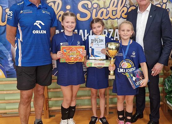 Dwóch dorosłych mężczyzn i trzy zawodniczki z pucharami i dyplomami pozują do zdjęcia w hali sportowej.