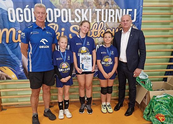 Dwóch dorosłych mężczyzn i trzy zawodniczki z pucharami i dyplomami pozują do zdjęcia w hali sportowej.