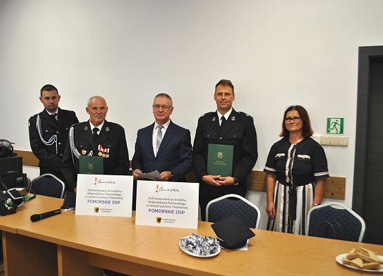 Przedstawiciele OSP, Wójt Gminy oraz przedstawicielka Urzędu Marszałkowskiego w gdańsku stoją z stołem prezydialnym, pozują do zdjęcia.