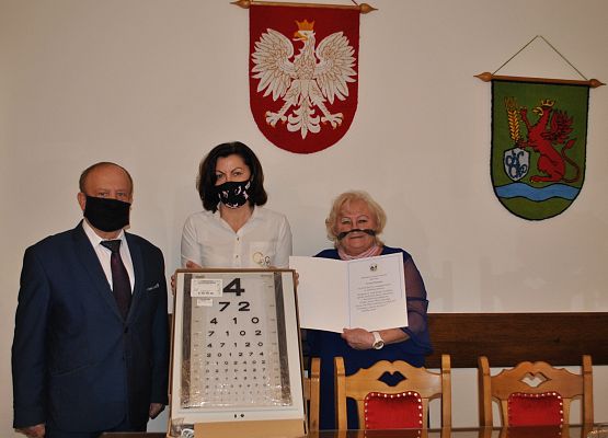 Gabinet wójta gminy. Od lewej wójt, dyrektor szkoły trzymająca tablicę fotometryczną, pielęgniarka szkolna trzymająca dyplom.