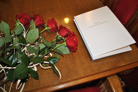 Pięć czerwonych róż leżących na stole obok białej teczki promocyjnej Gminy Nowa Wieś Lęborska.