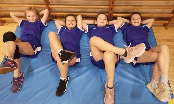 Cztery uśmiechnięte zawodniczki leża na materacu na hali sportowej i pozują do zdjęcia.