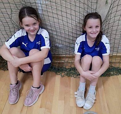 Dwie uśmiechnięte dziewczynki siedzą na parkiecie przy siatce w hali sportowej.
