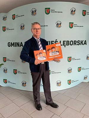 Wójt Gminy stoi na tle ścianki promocyjnej i trzyma książkę i segregator z materiałami projektu "ABC Empatii". grafika