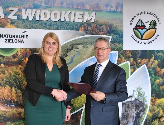 Na tle ścianki promocyjnej gminy wójt gminy wręcza swojemu zastępcy akt powołania. grafika