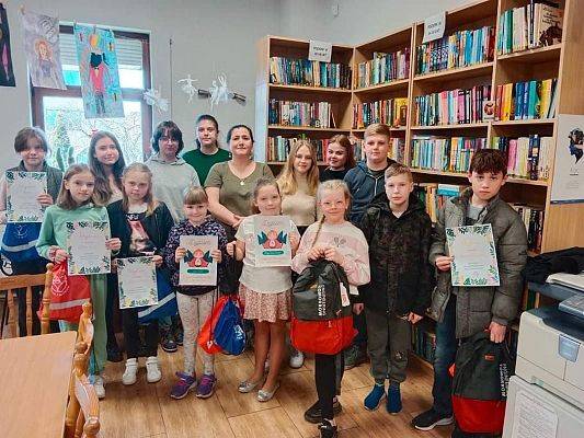 W sali biblioteki nagrodzeni uczestnicy konkursu z dyrektor biblioteki pozują do zdjęcia z dyplomami i nagrodami.