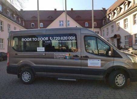 Samochód typu minibus, oznakowany do wykonywania usługi transportu door-to-door, na tle budynku Starostwa Powiatowego w Lęborku grafika