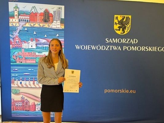 Laureatka stypendium stoi z dyplomem na tle ścianki promocyjnej województwa pomorskiego. grafika