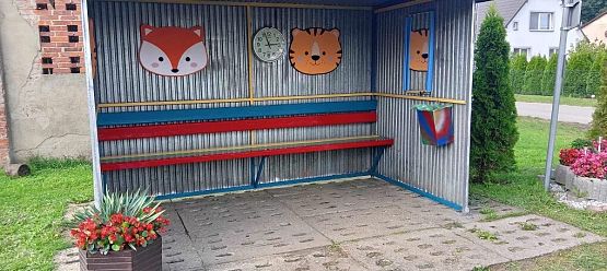 Blaszana, szara wiata przystankowa ozdobiona kolorowymi głowami zwierząt. Ławka pomalowana na kolorowo.