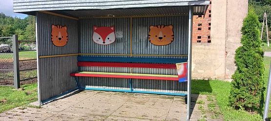 Blaszana, szara wiata przystankowa ozdobiona kolorowymi głowami zwierząt. Ławka pomalowana na kolorowo.