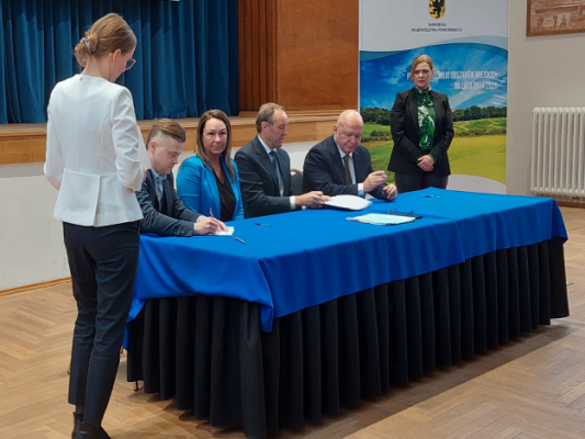 W sali konferencyjnej urzędu marszałkowskiego cztery osby siedzą za stołem prezydialnym i podpisują umowę.