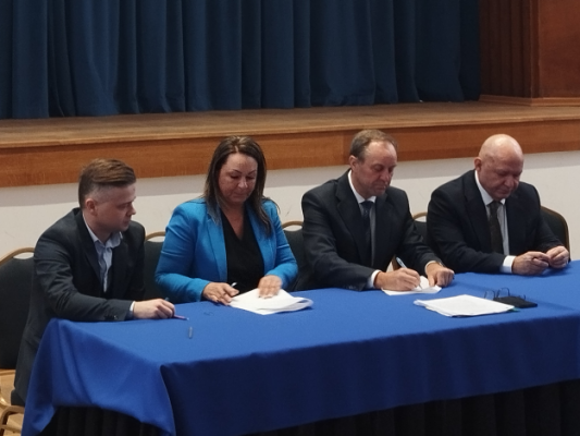 W sali konferencyjnej urzędu marszałkowskiego cztery osby siedzą za stołem prezydialnym i podpisują umowę.
